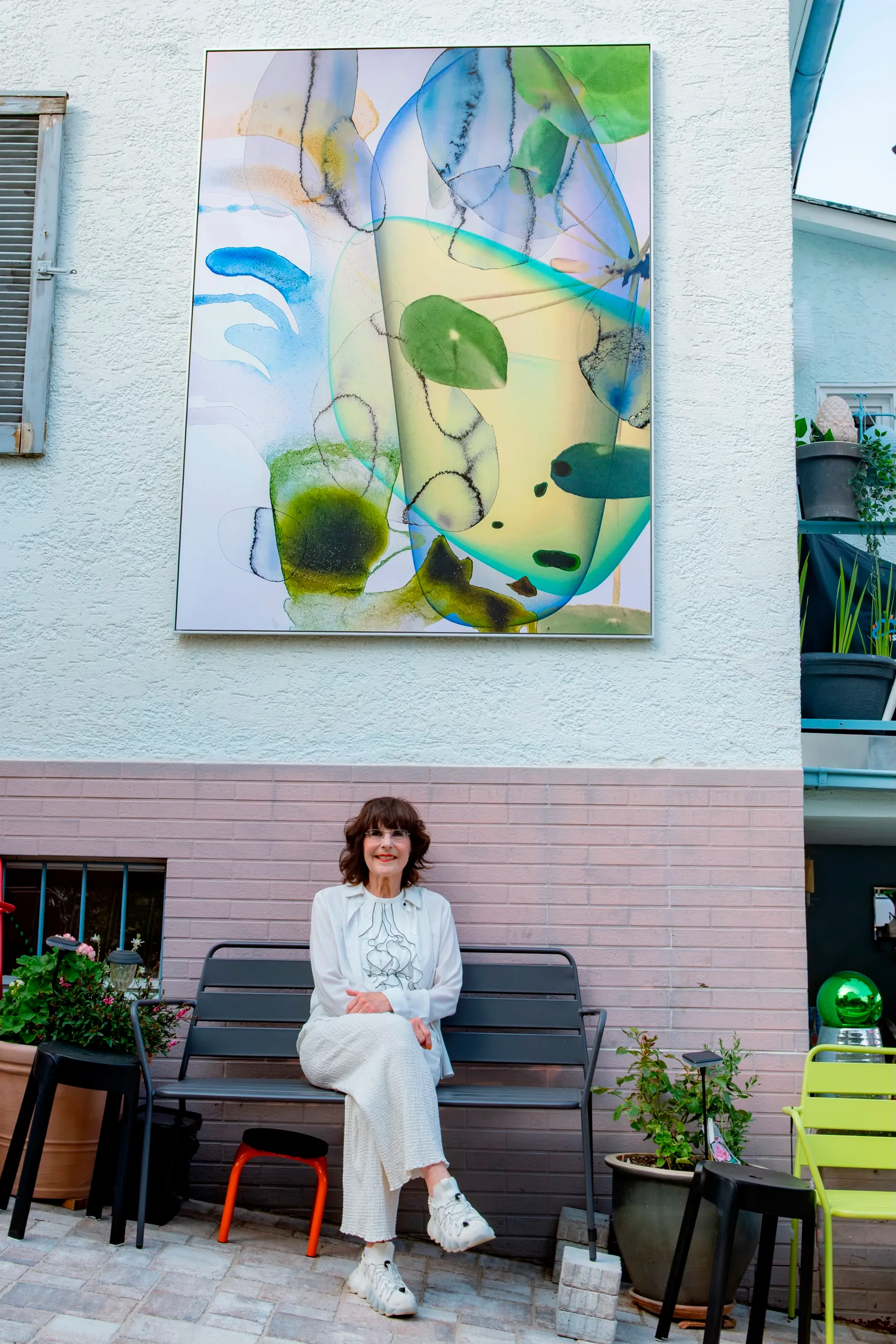 Christa Winter auf einer Bank vor ihrem Atelierhaus, über ihr ein großformatiges Werk
